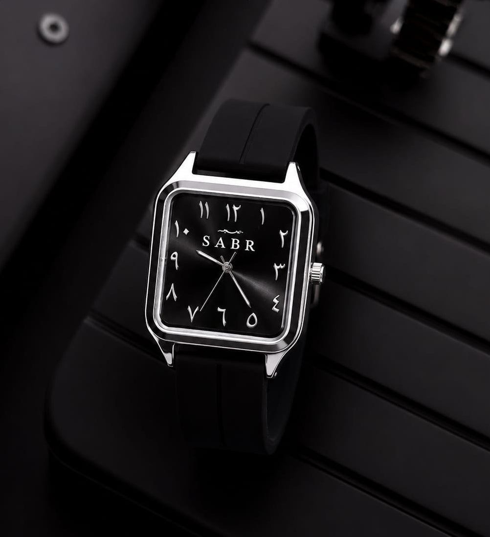 A square analog wristwatch with a black strap, silver-toned case, and black dial featuring white Arabic numerals and the brand name 'SABR'. The watch is displayed on a black slatted surface with a blurred second watch in the background.