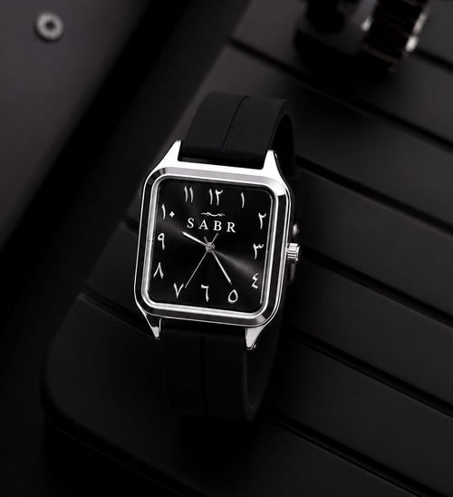 A square analog wristwatch with a black strap, silver-toned case, and black dial featuring white Arabic numerals and the brand name 'SABR'. The watch is displayed on a black slatted surface with a blurred second watch in the background.